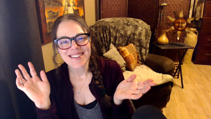 Femme avec lunette souriante assise à bureau avec devant elle un mirco. Elle pointe l'index de sa main gauche vers le haut.